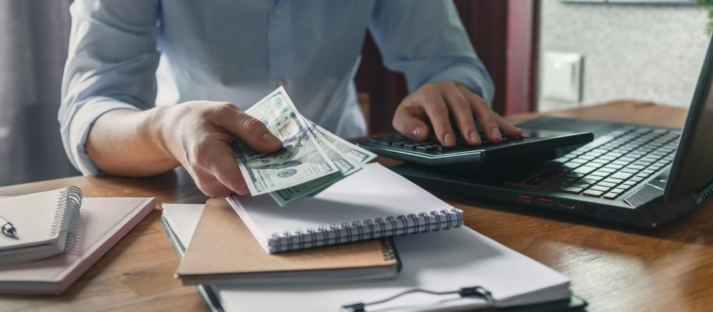 Home Budget Financial Planning In Crisis. Woman Quarantined And Working, Planning Home Budget, Lockdown Covid 19. Business Woman Holding A Pen And Press Calculator For Calculating Financial Report,