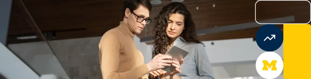 Two individuals using a tablet in a modern space with the University of Michigan logo